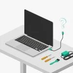 A laptop on a desk with an unplugged charging cable floating nearby, alongside a small repair toolkit and power adapter