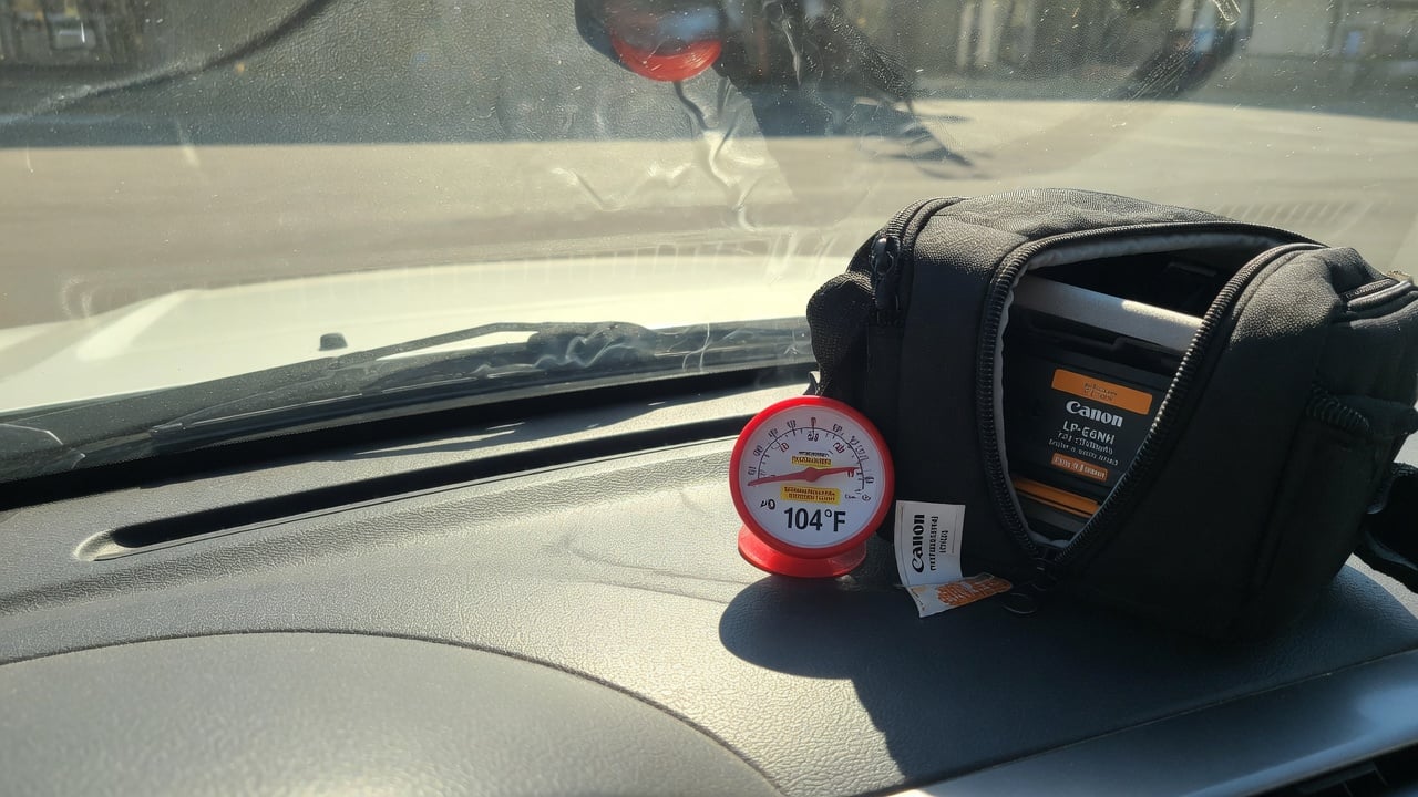 Black camera bag on a hot car dashboard with thermometer showing 40°C, illustrating heat damage risk to camera batteries