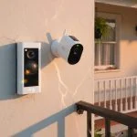 White video doorbell and wire-free security camera mounted on a sunlit house wall in direct afternoon sun