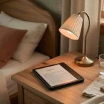 A Kindle-style e-reader resting on a wooden nightstand next to a lamp, showing weeks-long battery life in everyday use