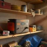 A Jackery-style portable power station sitting on a garage shelf, plugged into a wall charger with a small USB charge limiter device visible between the charger and the power station