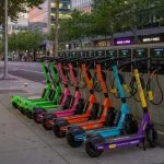 A row of brightly colored shared e-scooters lined up on a city sidewalk charging cabinet, each connected to a smart charger with cables visible