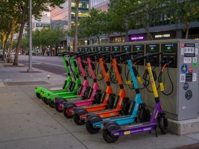 A row of brightly colored shared e-scooters lined up on a city sidewalk charging cabinet, each connected to a smart charger with cables visible