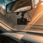 Compact dash cam mounted behind the rear-view mirror of a car, sunlight streaming through the windshield onto the dashboard