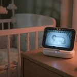 Video baby monitor parent unit sitting on its charging dock on a nightstand next to a crib, glowing screen
