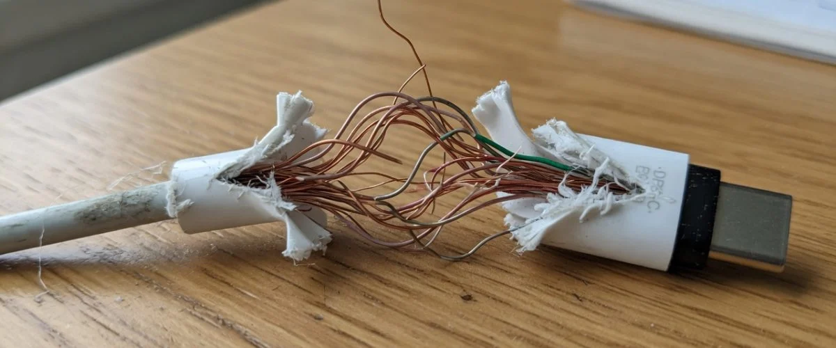 A frayed USB-C charging cable with exposed copper wires near the connector, resting on a wooden desk