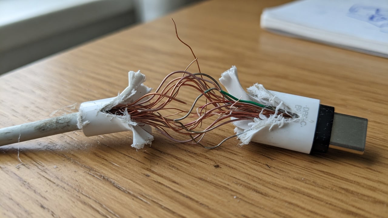 A frayed USB-C charging cable with exposed copper wires near the connector, resting on a wooden desk