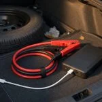 Compact lithium car jump starter pack in an open car trunk with jumper clamps and a USB-C cable plugged into its charging port